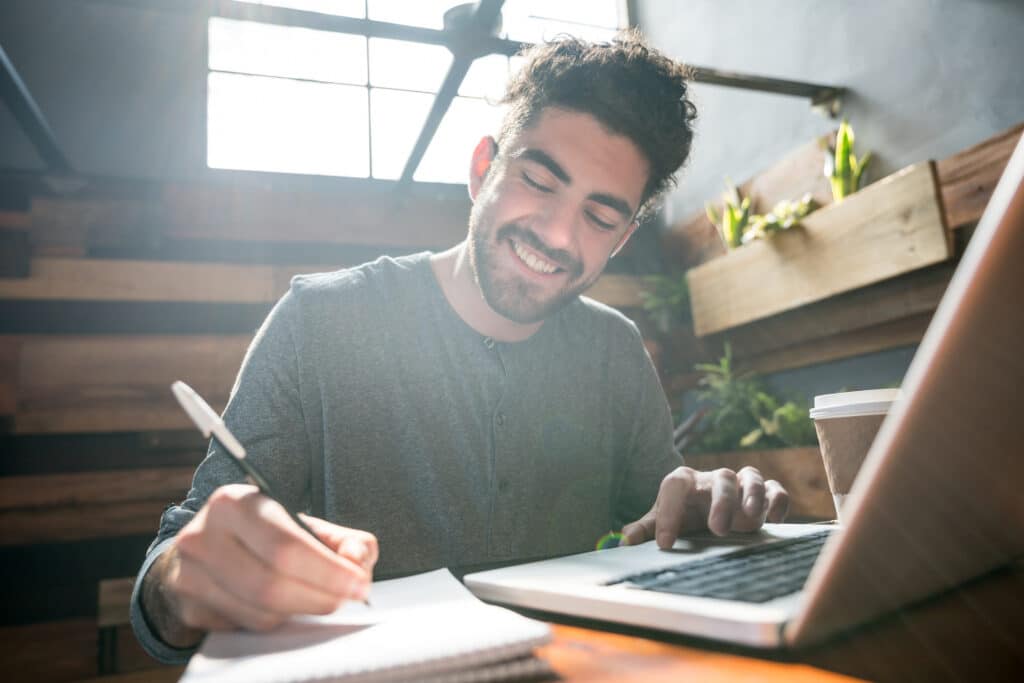 Empresario feliz en el restaurante de su pequeña empresa trabajando en un ordenador portátil y un bloc de notas sonriendo muy feliz mientras disfruta de un café.