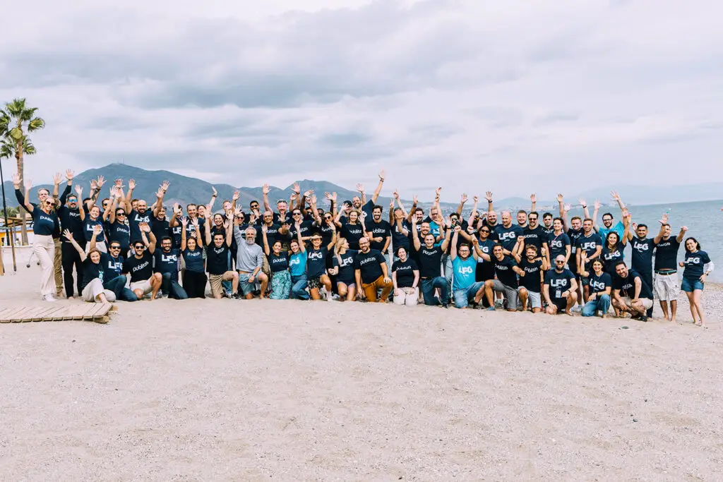 Publitas team. Huge group photo on the beach with people waving and smiling towards the camera.