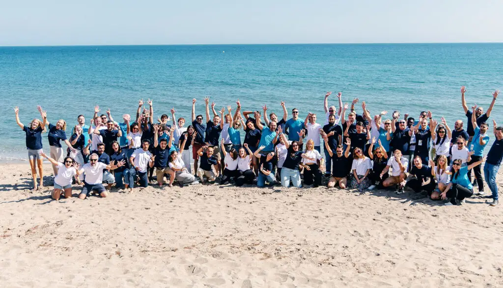 Publitas team. Huge group photo on the beach with people waving and smiling towards the camera.